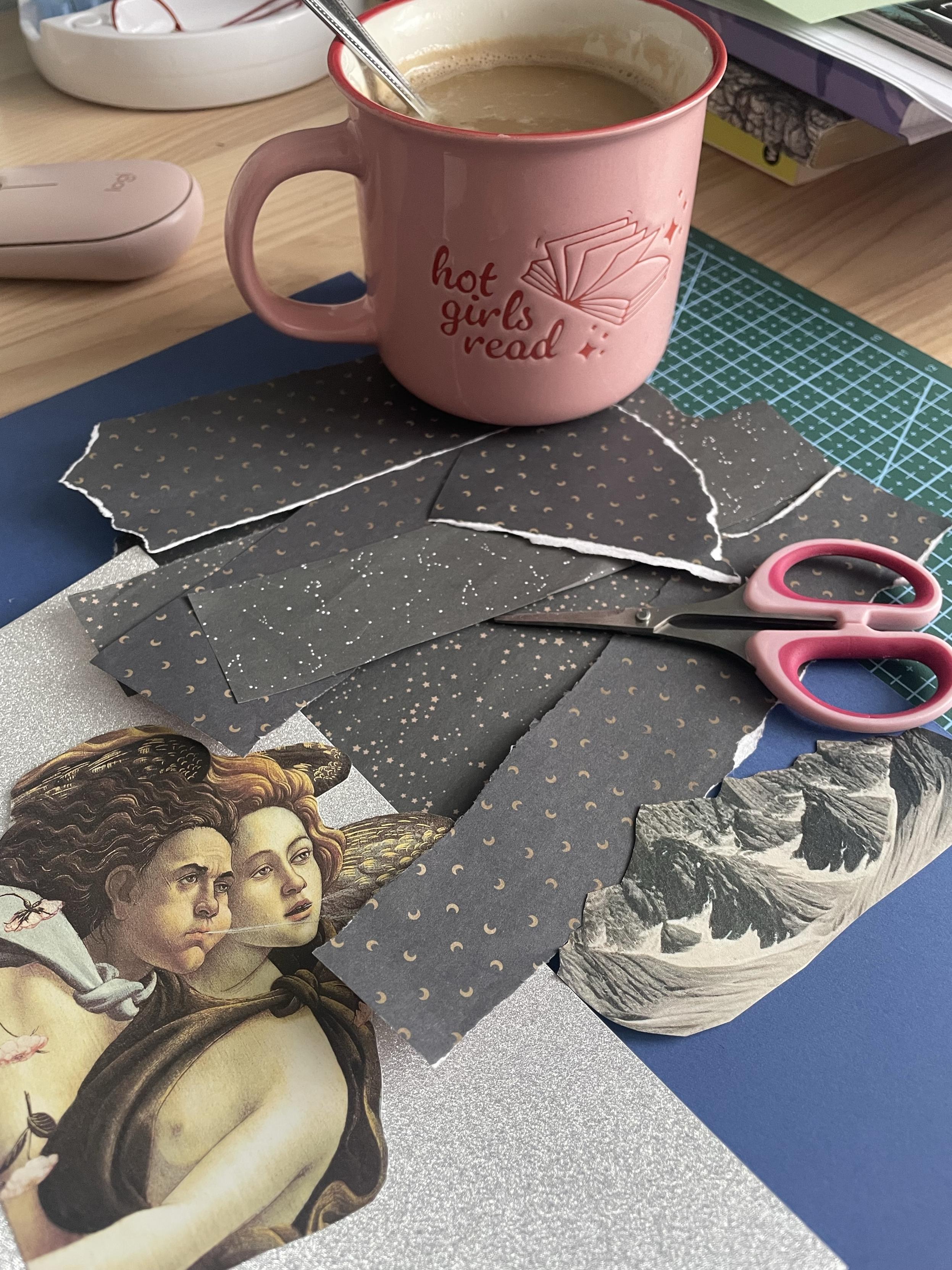A pink mug labeled "hot girls read" contains coffee, accompanied by a spoon. Surrounding the mug are various pieces of decorative paper in gray with patterns, scissors, and a collage element featuring a classical artwork of two figures. The items are placed on green pad.